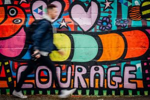 Wandbild in lebendigen, leuchtenden Farben um die Großbuchstaben "COURAGE". Ein Fußgänger läuft daran vorbei.